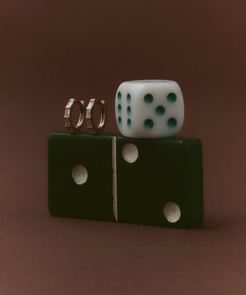 A green domino with two white dots stands upright on a brown surface. On top, a white die with five green dots and the Merit Baguette Hoops, made of recycled gold, are displayed. The background is plain brown.