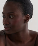 A woman with dark skin and smooth, pulled-back hair looks to the side, wearing a thin-strap top and delicate Merit Baguette Hoops. The background is plain and light-colored.