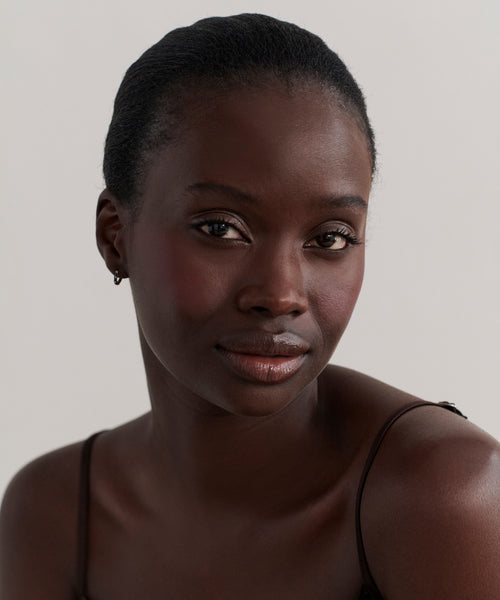 A woman with dark skin and short, pulled-back hair gazes at the camera with a neutral expression, wearing a thin strap top and delicate Merit Baguette Hoops in 14k gold against a light, plain background.