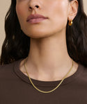 A woman wears the Merit Chain necklace, a 14k solid gold Cuban chain, along with a brown top and gold hoop earring. The image is cropped to show her face from the nose down and part of her upper chest.
