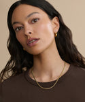 A woman with long wavy dark hair, wearing a brown top, gold hoop earrings, and two 14k solid gold Merit Chain necklaces, poses against a beige background. She looks slightly upward with a neutral expression.
