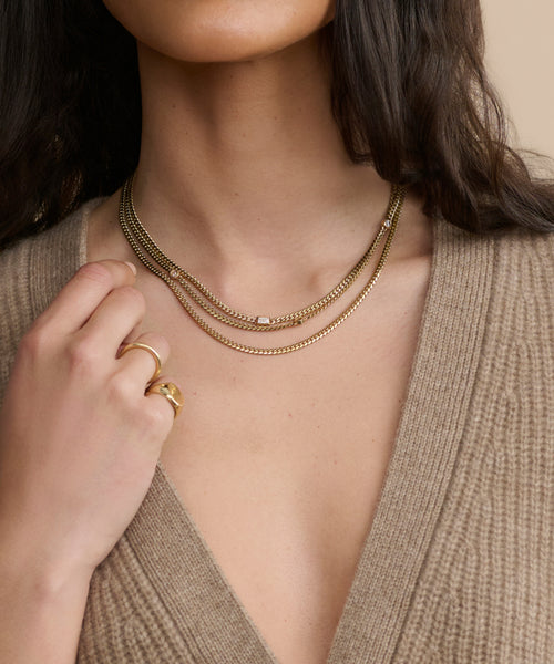 A woman, shown from the shoulders up in a beige ribbed deep V-neck sweater, wears layered gold necklaces including the Merit Emerald Cut Necklace and a gold ring, with long dark hair partially visible.