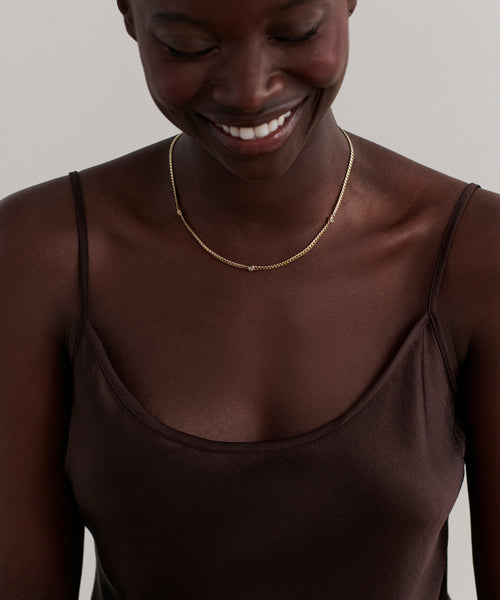A woman wearing a dark brown camisole smiles while looking down, showcasing the Merit Round Cut Diamond Necklace against a plain light background.