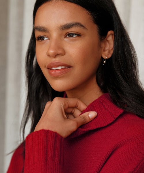 A woman with long dark hair wears a red knit sweater and delicately holds the collar, gazing to the side with a thoughtful look. She has natural makeup and completes her style with Mermaid Earrings featuring freshwater pearls.