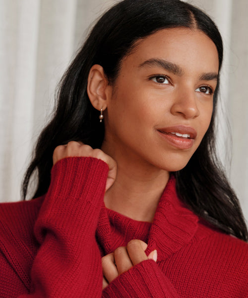 A woman with long dark hair, wearing a red knit sweater and smiling softly, holds her collar while elegant Mermaid Earrings with freshwater pearls glimmer gently.