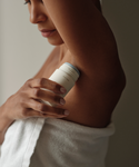 A person wrapped in a white towel applies Microbiome Balancing Deodorant, which is aluminum-free, to their underarm against a soft, neutral background.