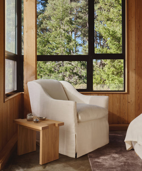 A cozy corner features the Miramar Chair—white Belgian linen, handmade in the USA—paired with a small wooden side table by large windows with views of green trees in a wood-paneled, naturally lit room.