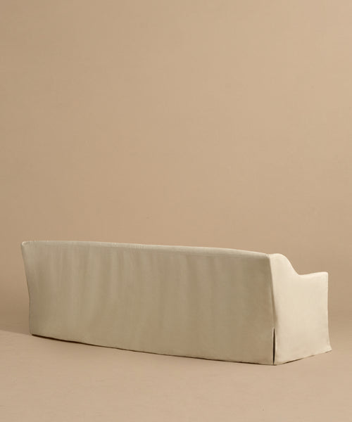 The Miramar Sofa, shown from the back against a plain light brown background, features a beige slipcover, clean lines, soft edges, and the inviting comfort of Belgian linen in a minimalistic design.