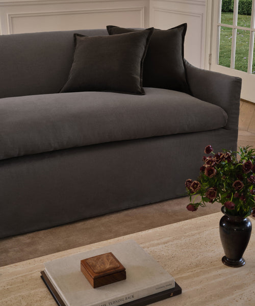The Miramar Sofa in dark grey Belgian linen with two matching cushions is placed against a white-paneled wall, while a coffee table holding books, a wooden box, and a vase of dark flowers stands in front. Sunlight enters through a glass door.