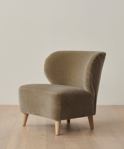 The Brentwood Chair, inspired by Jenni Kayne Furniture, features a modern armless design with a curved backrest, upholstered in light brown velvet, and stands on four light wood legs against an off-white wall.