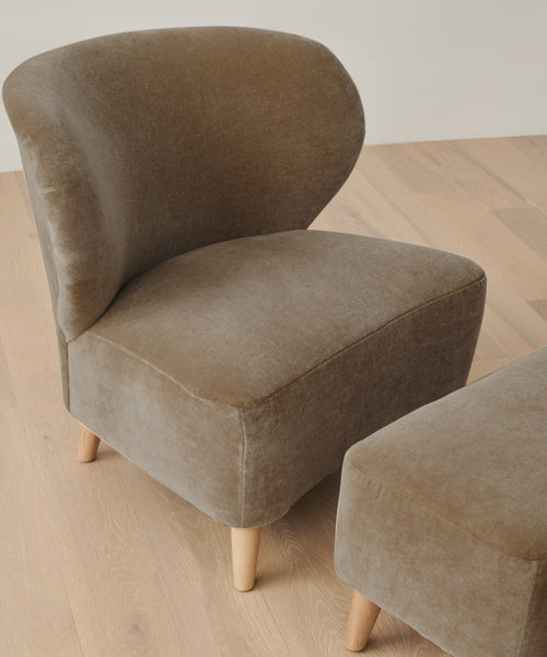 The Brentwood Chair features light brown velvet upholstery and light wooden legs, paired with a matching ottoman on a light wood floor, adding refined elegance to any space.