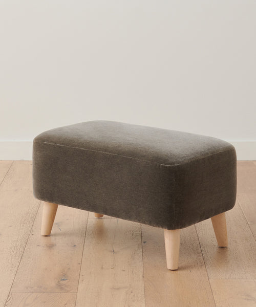 The Brentwood Ottoman features a small rectangular design with dark grey mohair upholstery and four light wooden legs, placed on a light wood floor against a plain white wall.