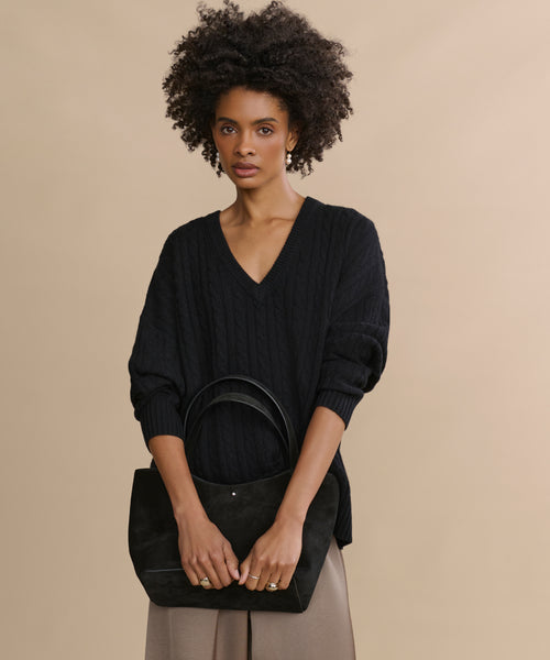 A woman with curly hair stands against a beige background, wearing the Nell V-Neck Sweater in black and taupe pants, holding a black handbag with both hands.