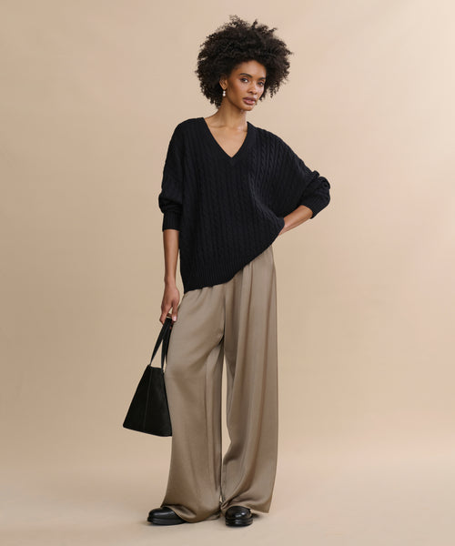A woman with short curly hair poses confidently against a beige background, wearing the Nell V-Neck Sweater in black, wide-legged beige trousers, black shoes, and holding a black handbag.