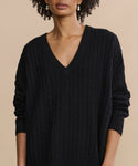 A woman wears the Nell V-Neck Sweater in black, standing against a beige background. Her curly hair and gold earrings are partially visible, complementing the cable knit design of the sweater.