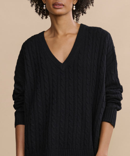 A woman wears the Nell V-Neck Sweater in black, standing against a beige background. Her curly hair and gold earrings are partially visible, complementing the cable knit design of the sweater.