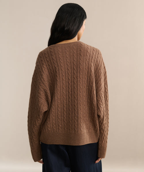 A person with long dark hair, seen from behind, wears the Nell V-Neck Sweater in brown and dark pants while standing against a neutral beige background.