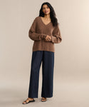 A woman with long dark hair stands against a neutral background wearing the Nell V-Neck Sweater, paired with wide-leg navy pants and black sandals. Her posture is relaxed and confident.