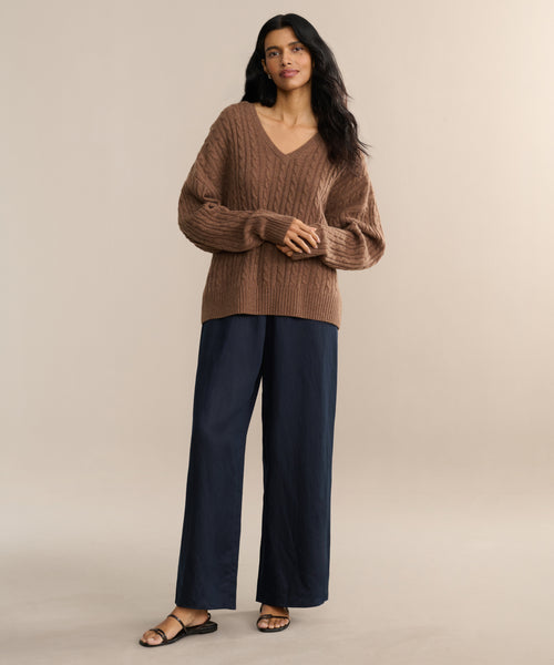 A woman with long dark hair stands against a neutral background wearing the Nell V-Neck Sweater, paired with wide-leg navy pants and black sandals. Her posture is relaxed and confident.
