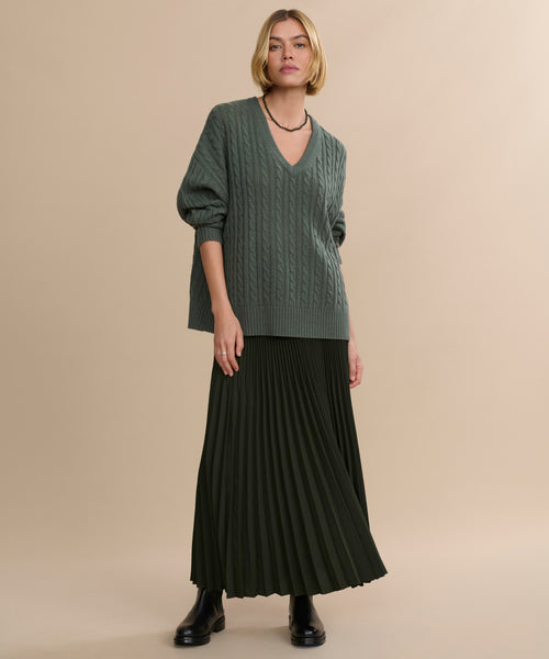 A woman with short blonde hair and a neutral expression stands against a beige background wearing the Nell V-Neck Sweater, a long dark green pleated skirt, black ankle boots, and a black necklace.