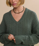 A woman with a chin-length blonde bob wears the green Nell V-Neck Sweater, a chunky relaxed fit cable knit with long sleeves, and a black beaded necklace. She stands against a neutral background, her face partially visible.