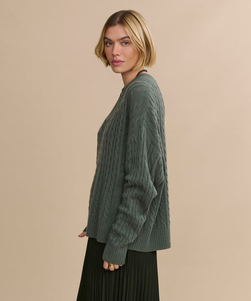 A person with short blonde hair wears the relaxed-fit Nell V-Neck Sweater in green and a pleated black skirt, standing sideways against a plain beige background.