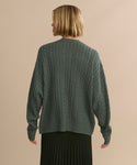 A person with short blonde hair, seen from behind, wears the Nell V-Neck Sweater in green and a pleated dark skirt, standing against a plain beige background.