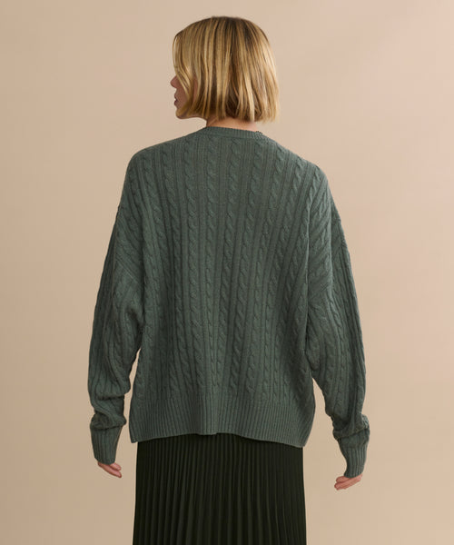 A person with short blonde hair, seen from behind, wears the Nell V-Neck Sweater in green and a pleated dark skirt, standing against a plain beige background.