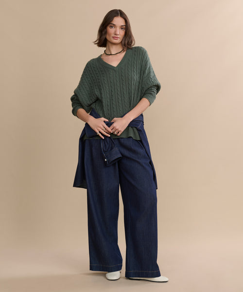 A woman stands against a beige background wearing the Nell V-Neck Sweater in green, wide-leg blue jeans, a blue jacket tied around her waist, white shoes, and a choker necklace.