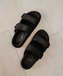 The Neoprene Hutton Slide Sandals in black feature two adjustable velcro straps per sandal and a molded footbed, shown here on a light wood floor.