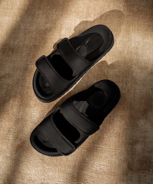 The Neoprene Hutton Slide Sandal in black features two wide, adjustable Velcro straps on each sandal, set against a textured beige surface with soft shadows.