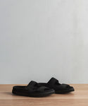 Two black Neoprene Hutton Slide Sandals with wide straps and molded footbeds for comfort are displayed side by side on a wooden surface against a light gray background.