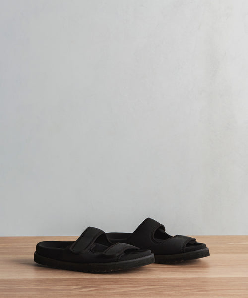 Two black Neoprene Hutton Slide Sandals with wide straps and molded footbeds for comfort are displayed side by side on a wooden surface against a light gray background.