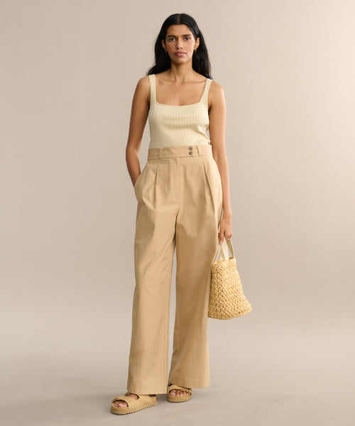 Against a plain beige background, a woman wears a cream tank top, high-waisted wide-leg Newport Trouser in khaki, beige platform sandals, and holds a woven straw handbag.