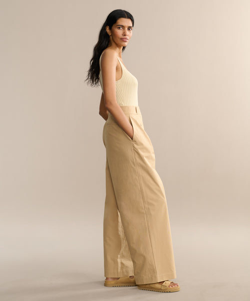 A woman with long dark hair stands sideways, hands in her pockets, wearing a sleeveless cream top, the Newport Trouser in khaki cotton poplin, and beige sandals against a plain beige background.