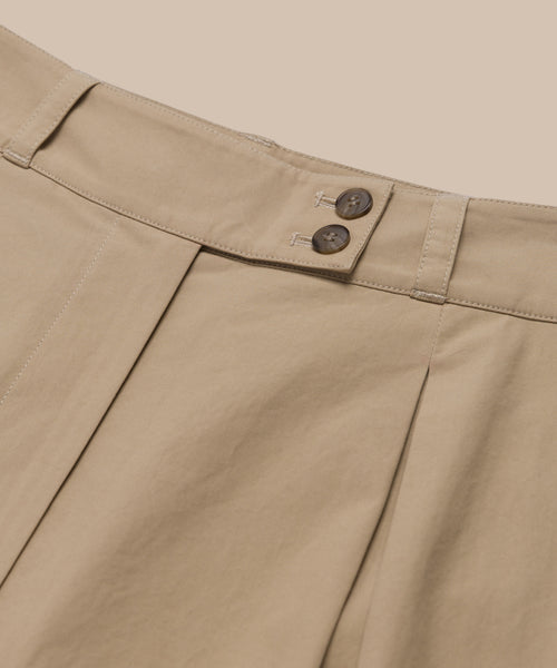 Close-up of the Newport Trouser in beige Italian cotton poplin, featuring pleats, a waistband with two dark brown buttons, and belt loops against a light beige background.