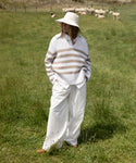 Wearing a wide-brimmed hat, striped sweater, white shirt, and relaxed Newport Trouser, a person stands in a grassy field while sheep graze in the background.