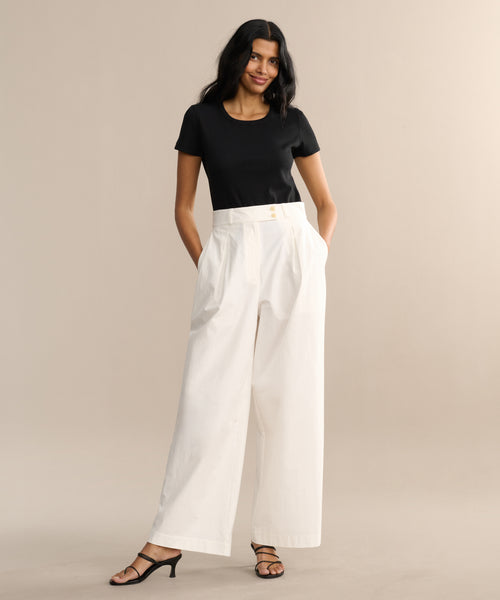 A woman stands with hands in her pockets against a beige background, wearing a fitted black short-sleeve top, the Newport Trouser, and black heeled sandals.