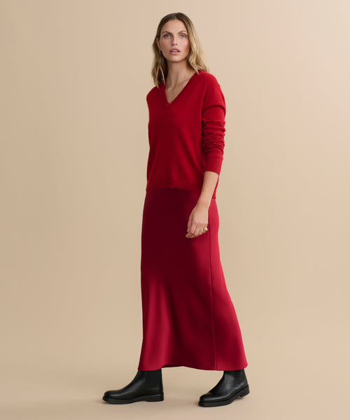 A woman stands against a beige background wearing the Nina Slip Skirt, a red V-neck sweater, and black ankle boots. She has loose, wavy blonde hair and gazes slightly to the side with a neutral expression.