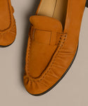 Close-up of two tan Nubuck Dylan Loafers with gathered toe stitching and a decorative upper strap, displayed on a light beige surface; expertly crafted in Italy.