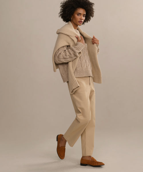 A woman poses in a chic, neutral outfit: a chunky cable-knit sweater, beige trousers, and elegant hand-stitched Nubuck Dylan Loafers in moccasin style, all set against a light background.