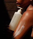 A person holds Awaken Body Wash, foam on their arm, bathed in warm sunlight against a dark wooden background.