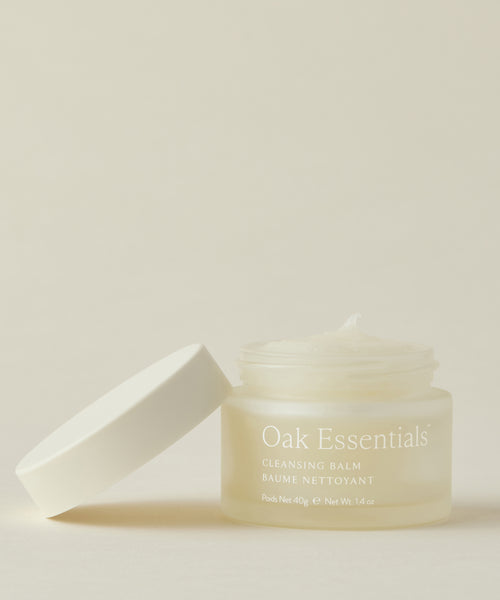 A frosted glass jar of Cleansing Balm with a white lid beside it, filled with a creamy balm-to-oil cleanser enriched with avocado oil, set against a light beige background.