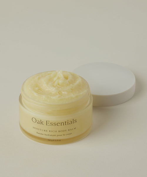 A glass jar of Moisture Rich Body Balm with a white lid sits on a light beige background, revealing its creamy, whipped texture enriched with cocoa butter inside.