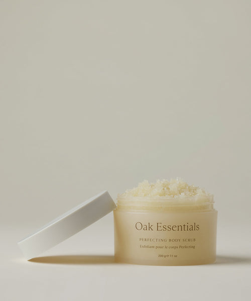A clear jar of Oak Essentials Perfecting Body Scrub, topped with a white lid, lies on its side. Inside is a grainy, light-colored exfoliant, displayed against a neutral off-white background.