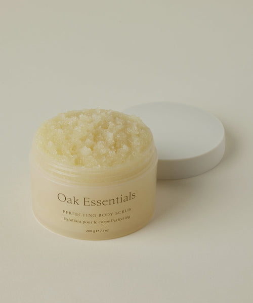 An open jar of Perfecting Body Scrub with its white lid beside it shows the textured, grainy exfoliating formula inside. Set on a neutral background, this hydrating skincare treat is beautifully highlighted.