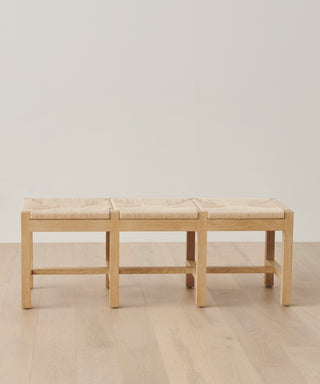 The Montecito Bench, featuring three woven Danish paper cord seats, sits on a light wood floor against a plain off-white wall.