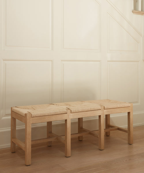 The Montecito Bench in light white oak with a woven Danish paper cord seat is placed by a cream paneled wall near the stairs on a wooden floor.