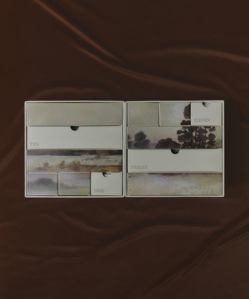 Two Oak Essentials x Jenni Kayne Advent Calendar boxes, each featuring twelve numbered rectangular compartments and soft landscape art, are displayed on a brown silky fabric background.