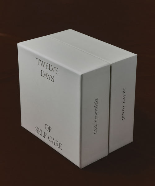 A white rectangular box with minimalist text reads Twelve Days of Self Care. The sides show Oak Essentials x Jenni Kayne, creating a chic Advent Calendar perfect for self-care gifting, set against a dark brown background.